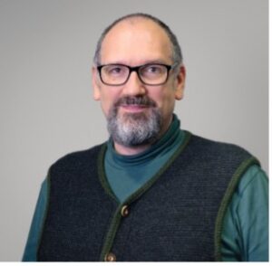 Middle-aged man with a short beard, glasses, and a green turtleneck under a dark gray vest, smiling against a neutral gray background.