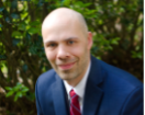 Young white man in blue suit, white shirt, and red tie in front of some plants